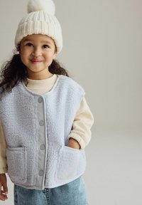 Lichtblauw fleece vest met een gestructureerde stof, grijze accenten en twee voorzakken, gedragen over een crèmekleurige trui en gecombineerd met een witte gebreide muts.