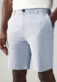 Pantalones cortos de hombre en tela a rayas blancas y azul claro, con cierre de botón, bolsillos laterales y un corte ajustado. Textura suave con un acabado liso.