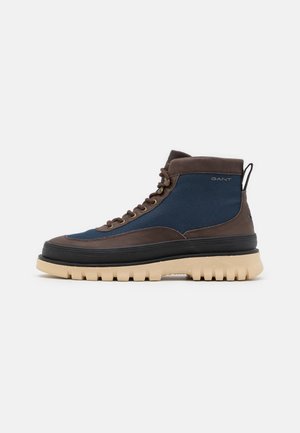 Wysokie casual buty z niebieskiej tkaniny z brązowymi skórzanymi wykończeniami, czarną środkową podeszwą, beżową masywną podeszwą i logo po boku na jasnoszarym tle.