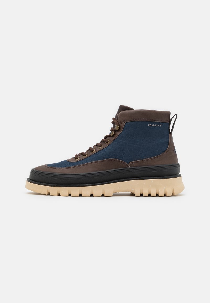Botte décontractée montante en tissu bleu avec garniture en cuir marron, semelle intermédiaire noire, semelle épaisse beige et logo latéral sur fond gris clair.