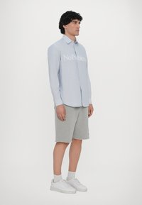 Cămașă bleu deschis, cu butoni, inscripționată cu textul "No Problem", purtată cu pantaloni scurți gri și adidași albi. Materialul pare neted, cu o ajustare ușor structurată.