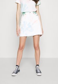 Mujer vestida con una falda blanca y de colores pastel con dobladillo sin terminar, blusa a juego y zapatillas altas oscuras de color azul, de pie contra un fondo liso.