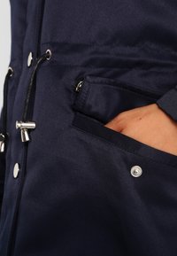 Ruka částečně uvnitř kapsy tmavě modré bundy se stříbrným patentem a detaily černého stahovacího táhla.