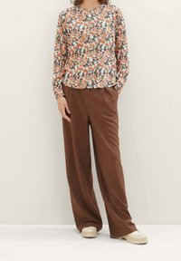 Blusa floreale a maniche lunghe con motivi arancioni, blu e verdi; abbinata a pantaloni marroni larghi e scarpe di colore chiaro.