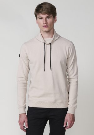 Joven vestido con una sudadera texturizada de un beige claro con cordones negros y pantalones negros, de pie frente a un fondo gris liso.