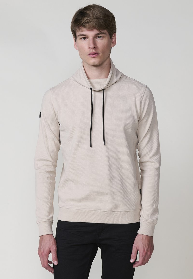 Giovane uomo che indossa un hoodie beige chiaro a trama con lacci neri e pantaloni neri, in piedi di fronte a uno sfondo grigio uniforme.