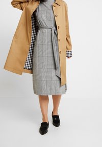 Abrigo trench beige sobre un vestido de cuadros en gris y blanco con cinturón en la cintura. Unos mocasines negros completan el conjunto. El forro a cuadros es visible.