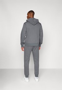 Grijze hoodie met een buidelzak en capuchon, gecombineerd met bijpassende grijze sweatpants. Beide items hebben geribbelde manchetten en een zachte textuur. Witte sneakers.