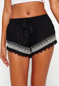 Shorts noirs à coupe décontractée, ornés de motifs brodés blancs et d'une dentelle au bas. Taille réglable avec des pompons noirs.