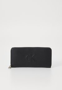 Portefeuille zippé noir en cuir lisse, avec un logo "cK" en relief à l'avant et une forme rectangulaire élégante.