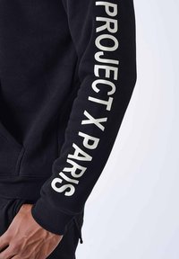 Sudadera negra con gran texto blanco en la manga que dice 'PROJECT X PARIS', con puños acanalados y cordón en el dobladillo.