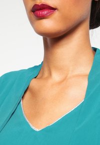 Vêtement en teal avec un col en V et des bordures en argent ; texture lisse, gros plan sur le cou et les lèvres avec un rouge à lèvres rouge profond, mettant en valeur le contraste des couleurs.