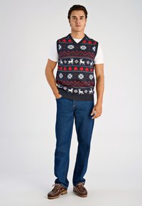 Suéter sin mangas azul marino con diseño estampado en rojo y blanco que presenta renos y copos de nieve, combinado con jeans azules y zapatos marrones.