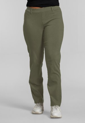 Person trägt olivgrüne, gerade geschnittene Hosen und weiße Turnschuhe, steht vor einem schlichten grauen Hintergrund.