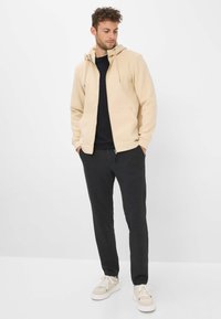 Felpa beige con zip e cordini, abbinata a pantaloni della tuta grigio scuro e sneakers chiare. Tessuto morbido; vestibilità casual e confortevole.