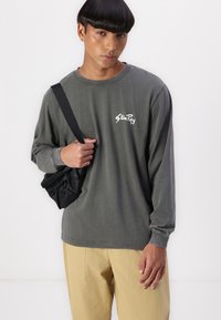 Long-sleeve gray cotton shirt featuring a white logo. Worn with beige trousers, model holds a black bag against a white background.