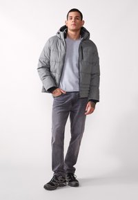 Grå polstret jakke med hette, med glidelås foran og sidelommer, brukt over en lys grå genser og mørke grå jeans med svarte joggesko.