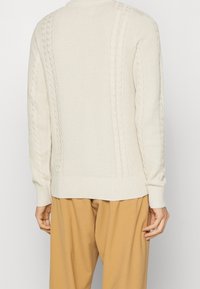 Maglione a maglia crema con motivi a treccia lungo i lati, polsini e orlo a costine, e scollatura rotonda. Indossato con pantaloni beige.