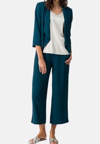 Lekki blezer w kolorze morskim z drapowanym przodem; kremowa, luźna bluzka; szerokie spodnie w kolorze morskim; sandały na metalicznych paskach. Gładkie tekstury w całej stylizacji.