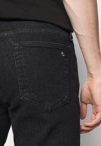 Jeans de mezclilla negra con un bolsillo trasero que presenta un discreto logo blanco. Los jeans tienen un acabado texturizado y un diseño clásico de cinco bolsillos.