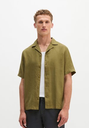 Man draagt een olijfgroen kort mouwen overhemd met knopen over een wit tanktop en zwarte broek, staand tegen een effen witte achtergrond.