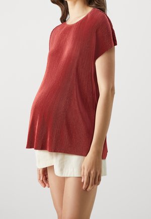Vrouw draagt een losse, getextureerde rode korte mouwen top over witte geribbelde shorts, staand tegen een effen lichte achtergrond.