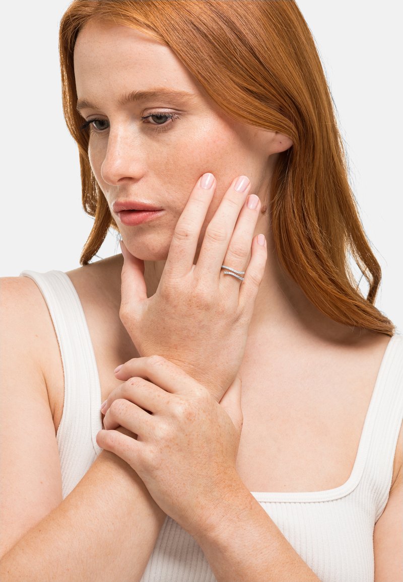 Sølvring med to sammenflettede bånd, prydet med små klare edelstener. Modellen har langt, stritt rødt hår og lys hud, iført en hvit tanktop.