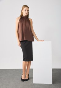 Bruine mouwloze top met hoge hals en geplooid ontwerp, gecombineerd met een strakke zwarte pencilrok en zwarte stilettohakken. Witte voetstuk in de buurt.