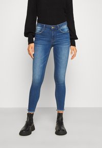 Jeans skinny albastre cu o spălare decolorată, cinci buzunare, închidere cu nasture și tiv raw la glezne, asortați cu un pulover negru tricotat.