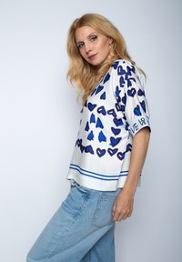 Weißes Blüschen mit blauen Herzmustern, kurzen Ärmeln und einem blauen Horizontalstreifen am Saum. Lockerer Schnitt mit dem Text "LOVE" auf dem Ärmel.