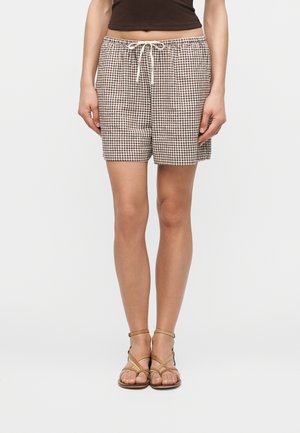 Άτομο που φοράει καφέ και λευκά καρό σορτς με κορδόνι στη μέση, σκούρο καφέ κοντό τοπ και ταμπά σανδάλια με λουριά.