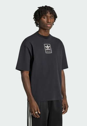 Młody mężczyzna z krótkimi dredami, ubrany w luźną czarną koszulkę Adidas i czarne spodnie z białymi paskami, stojący na jednolitym tle.
