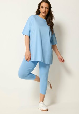 Woman standing on one leg wearing light blue oversized t-shirt, matching cropped leggings, and white sneakers against a plain background.