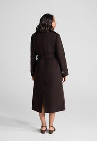 Cappotto lungo marrone con vita cintata, caratterizzato da polsini con bottoni e una fessura posteriore per facilitare i movimenti. Tessuto morbido, design su misura.
