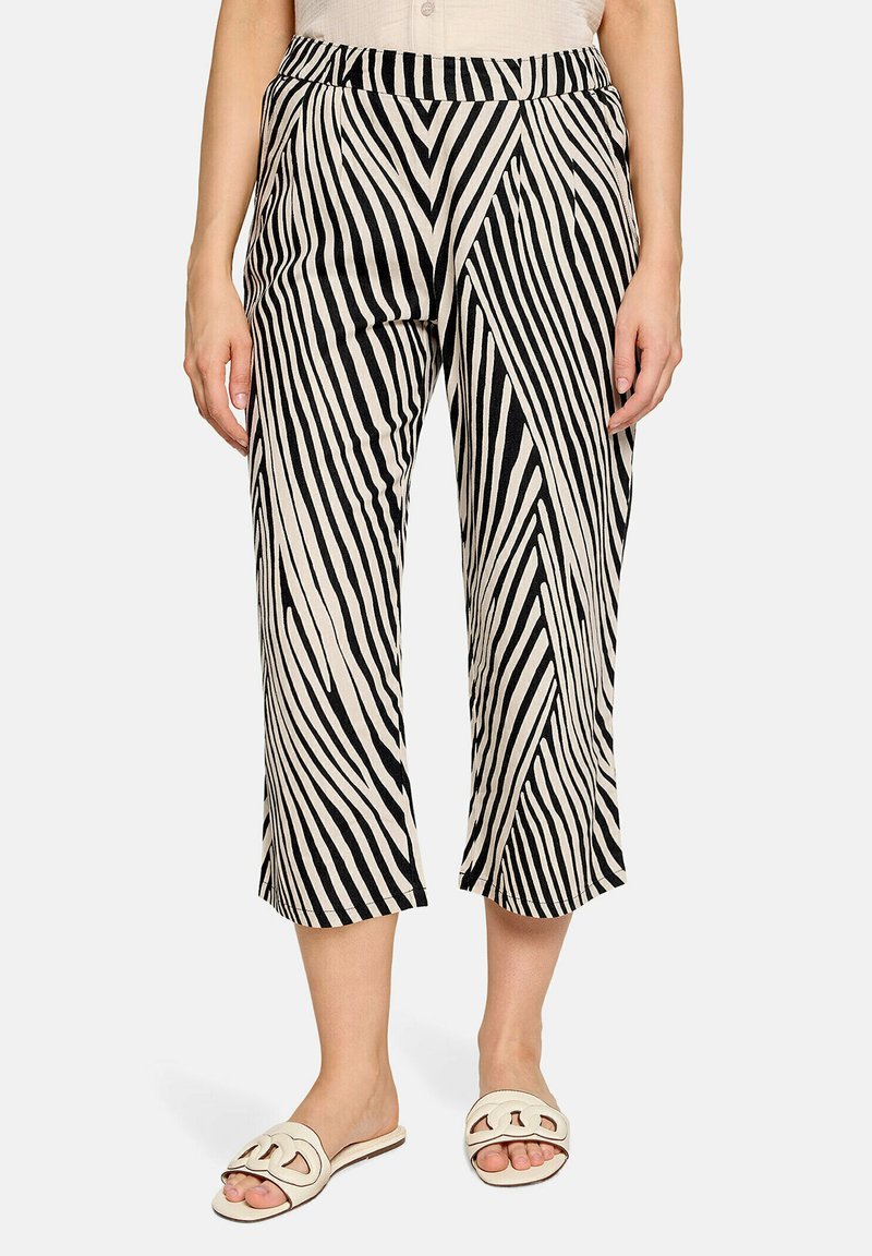 Femme portant un pantalon court avec un motif rayé diagonal noir et blanc et des sandales blanches ouvertes, debout devant un fond uni.