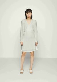 Vestido branco texturizado com mangas compridas, decote em V e cintura ajustada. Usado com sandálias de cor clara, contra um fundo simples.