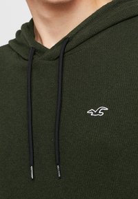 Mörkgrön hoodie i strukturerad tyg med svarta dragsnören. Har en vit broderad logotyp på bröstet.