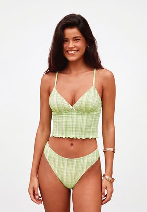 Uśmiechnięta kobieta z długimi ciemnymi włosami, ubrana w zielony kraciasty crop top i dopasowane majtki bikini z wysokim wycięciem, stojąca na jednolitym tle.