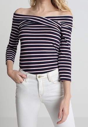 Frau trägt ein marineblaues schulterfreies Oberteil mit weißen und roten horizontalen Streifen und weiße hoch taillierte Hosen, Hand in der Tasche.