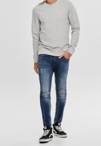 Felpa grigia a maniche lunghe, jeans slim-fit blu con lievi segni di usura e sneakers alte nere con lacci bianchi e logo.