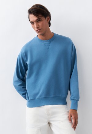 Mężczyzna o ciemnych włosach, ubrany w niebieską bluzę z długim rękawem i białe spodnie, stojący na tle gładkiego, jasnego tła.