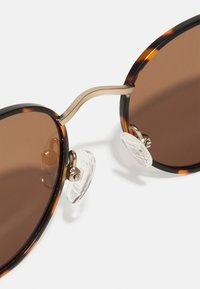 Ochelari de soare din material tortoiseshell cu lentile de culoare maro. Au un pod metalic auriu și pernuțe nazale transparente pentru confort. Formă rotundă și design ușor.