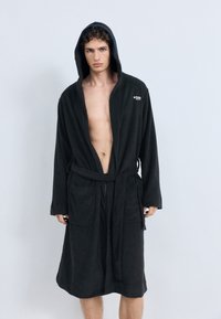 Robe de banho em tecido felpudo preto com capuz, mangas compridas, bolsos frontais e cinto para amarrar. Apresenta um pequeno logótipo na parte superior esquerda.