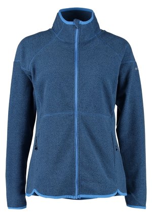 Mujer con chaqueta de forro polar azul con cremallera, vaqueros pitillo azules desgastados y zapatillas grises, de pie con una mano en el bolsillo.