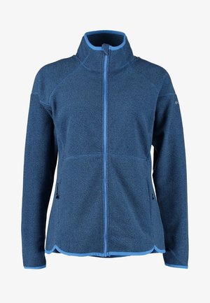 Mujer con chaqueta de forro polar azul con cremallera, vaqueros pitillo azules desgastados y zapatillas grises, de pie con una mano en el bolsillo.