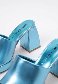 Mules en métal bleu avec un talon carré épais. Texture lisse, design minimaliste et accent doré à la base du talon.