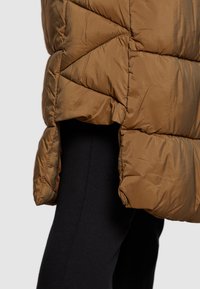 Brązowa kurtka puchowa o pikowanym wzorze, z wysokim kołnierzem i bocznymi rozcięciami dla łatwości ruchu, w połączeniu z czarnymi legginsami.