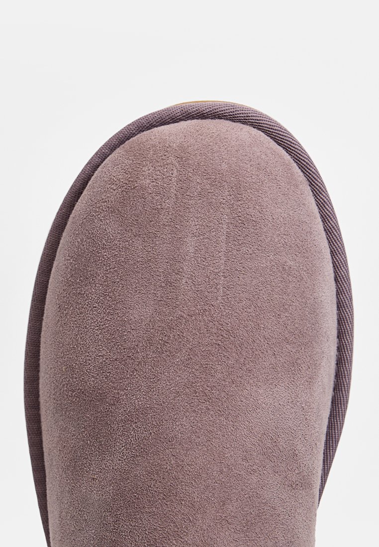 Pantofole in suede viola con punta arrotondata, texture morbida e una banda in tessuto a coste contrastante lungo il bordo.