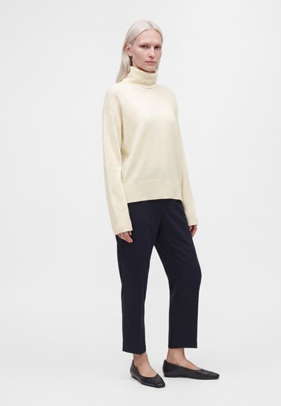Femme debout portant un pull à col roulé crème, un pantalon noir raccourci et des chaussures plates noires sur un fond blanc uni.