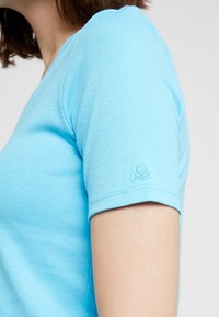 Vaaleansininen puuvillapaita, jossa on muotoonleikattu malli, lyhyet hihat ja pieni kirjailtu logo hihassa. Sileä pinta.